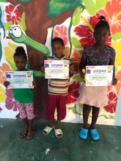 Three children holding up their certificates in front of a wall.