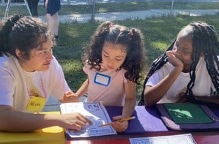 Three children sitting at a table with papers and pens.